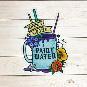Op de afbeelding: Een blauwe mok met de tekst "Don't Drink Paint Water" en een paarse rand. De mok heeft twee kwasten die eruit steken en is omringd door bloemen.