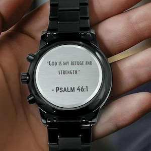 May include: Black wristwatch with a black metal band and a round silver face. The watch face has the text "God is my refuge and strength." - Psalm 46:1.
