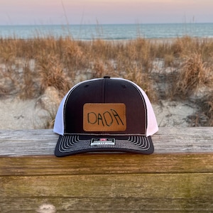 May include: Black and white trucker hat with a brown leather patch that says "DADA" on it. The hat is sitting on a wooden surface.
