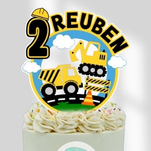 May include: A white cake with yellow and black construction vehicle themed frosting and a topper that says "Reuben 2" with a construction worker hat. The cake is decorated with colourful sprinkles and a small blue and pink camper sticker on the side that says "My Crafty Caravan".