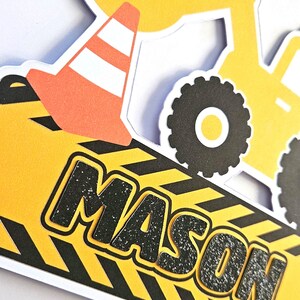 K&ouml;nnte beinhalten: Gelber Baufahrzeug-Kuchentopper mit einer schwarzen Zahl 3, einem schwarz-gelb gestreiften Schild mit dem Namen Mason und einem schwarz-orangenen Verkehrskegel.