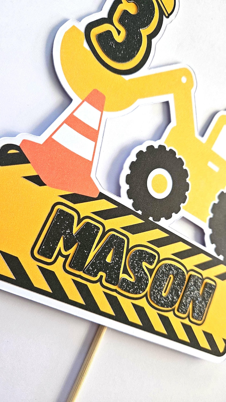 K&ouml;nnte beinhalten: Gelber Baufahrzeug-Kuchentopper mit einer schwarzen Zahl 3, einem schwarz-gelb gestreiften Schild mit dem Namen Mason und einem schwarz-orangenen Verkehrskegel.