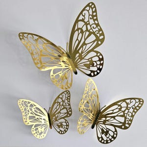May include: Three gold-coloured metal butterfly wall decorations. The butterflies have intricate cut-out wing designs and are in various sizes. The butterflies are attached to a white wall.
