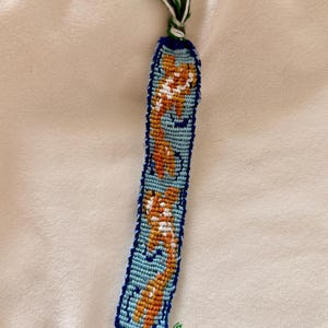 May include: Handmade friendship bracelet featuring a repeating orange and white design on a light blue background. The bracelet is bordered with dark blue thread and has a tassel of green, white, and black threads at the top.