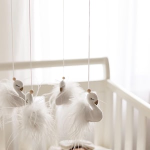 May include: A baby mobile with five white swan decorations, each with fluffy feather detailing, hanging from a wooden frame. The swans have small wooden bead heads, suspended above a white cot.