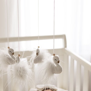 May include: A white baby mobile with four swan-shaped ornaments hanging from a wooden frame. The swans have white feathers and gold accents. The mobile is hanging above a white cot.