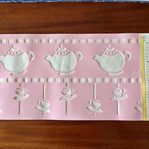 May include: Pink stencil featuring white teapots and teacups. The stencil includes the text "ME" and "Mary Engelbreit" and is designed for craft projects. The stencil is pre-cut and made by Delta.