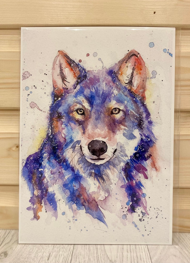 May include: Watercolour painting of a wolf's face in shades of blue, purple and brown. The wolf has a gentle expression and is surrounded by white splashes of paint.