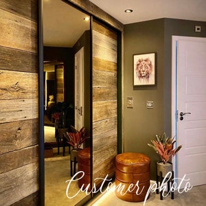 May include: A hallway with a sliding door made of reclaimed wood panels. The door has a mirror on one side. There are two leather ottomans and two potted plants in the hallway. The wall is painted a dark green color. A painting of a lion's face hangs on the wall. The text "Customer photo" is written on the floor.