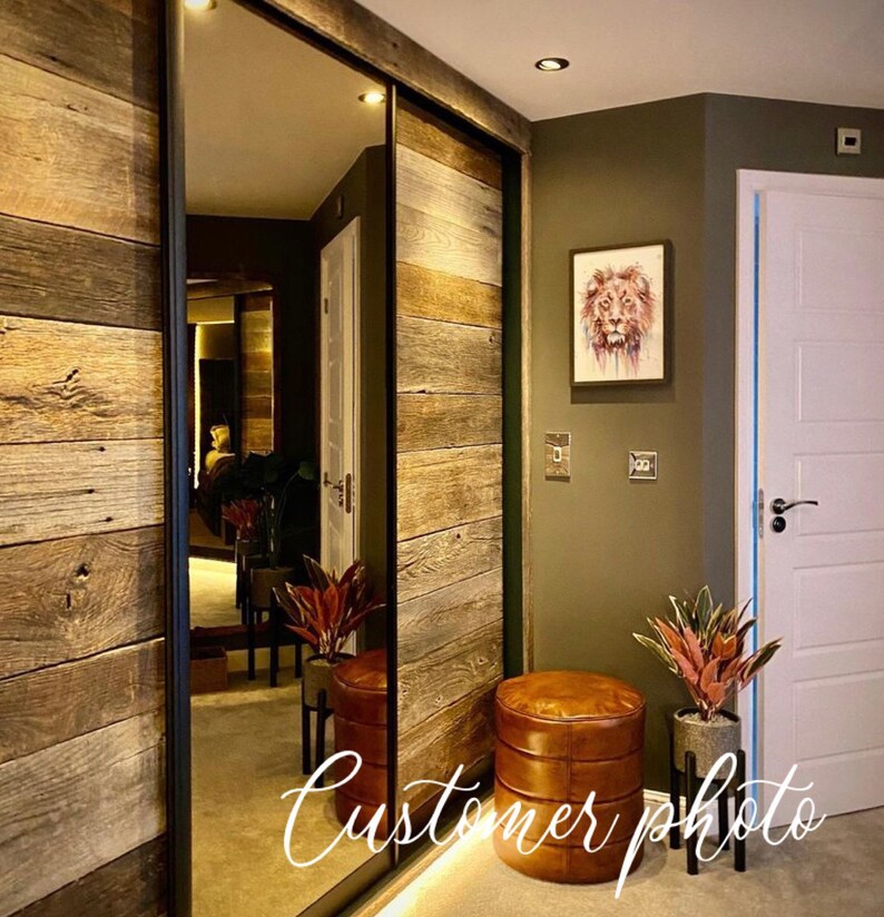 May include: A hallway with a sliding door made of reclaimed wood panels. The door has a mirror on one side. There are two leather ottomans and two potted plants in the hallway. The wall is painted a dark green color. A painting of a lion's face hangs on the wall. The text "Customer photo" is written on the floor.