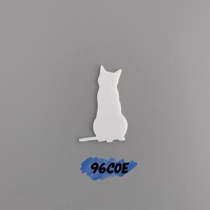 May include: White ceramic silhouette of a cat sitting on a gray background. The text "96COE" is written in blue paint on the background.