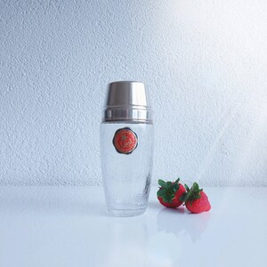 Può includere: Uno shaker per cocktail in vetro trasparente con un tappo argentato e un'etichetta rossa e dorata che dice "100 Jahre -  " e "Qualitat". Due fragole rosse sono in primo piano.