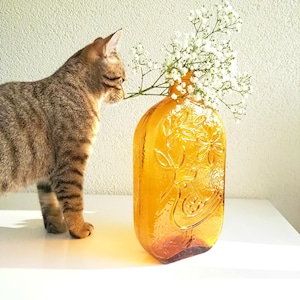 Op de afbeelding: Een tabbykat die aan een bruine glazen vaas met een bloemenmotief en witte baby's breath bloemen snuffelt.