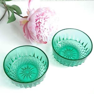 Op de afbeelding: Twee groene glazen kommen met een diamantpatroon en een stervormig ontwerp in het midden. De kommen staan op een wit oppervlak.