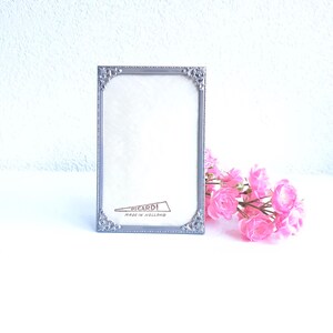 Può includere: Una cornice rettangolare argentata con dettagli ornati, con la scritta "PICARDI MADE IN HOLLAND". Fiori rosa sono disposti a destra della cornice. La cornice è vuota, pronta per una foto.