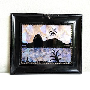 May include: Framed artwork with a black frame, showing a landscape scene. The artwork features a silhouetted mountain, palm trees, and a body of water. The sky has an iridescent pattern with white, purple, and gold hues.