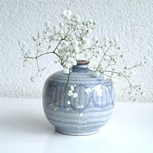 Puede incluir: Un jarrón pequeño, redondo, de cerámica azul y blanca con un arreglo de flores blancas. El jarrón tiene un patrón texturizado a rayas.