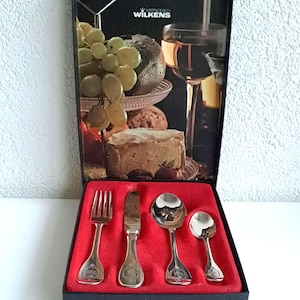 Könnte beinhalten: Ein schwarzes Etui mit roter Samt-Auskleidung, das ein Set aus vier versilberten Besteckteilen enthält. Die Box zeigt ein gedrucktes Bild von Trauben, Käse und einem Glas Wein. Das Besteck umfasst eine Gabel, ein Messer, einen Suppenlöffel und einen Teelöffel. Auf der Oberseite der Box steht "Wert & Design Wilkens" gedruckt.