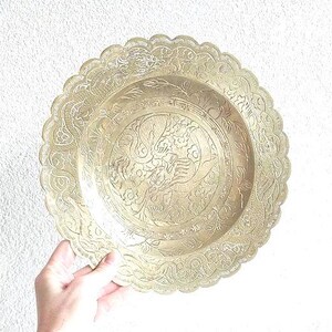 Op de afbeelding: Een ronde, messing serveerschaal met een ingewikkeld, gegraveerd ontwerp van een vogel en bloemmotieven. De schaal heeft een gegolfde rand.