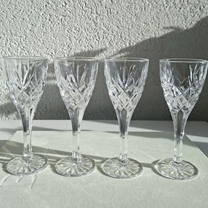 Op de afbeelding: Vier heldere kristallen wijnglazen met diamantgeslepen patronen. De glazen hebben lange stelen en cirkelvormige bases. De achtergrond is een witte muur met textuur en een schaduw.