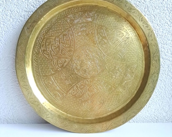 May include: A round, golden brass tray with intricate etched patterns. The tray features detailed floral and geometric designs, with a central medallion and a decorative border. The brass has a warm, polished finish, reflecting light. This decorative tray is suitable for serving or display.