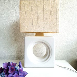 May include: A table lamp featuring a white square ceramic base with a large circular cutout in the centre. The lamp is topped with a rectangular beige lampshade that has a textured, fibrous surface with vertical line details throughout.