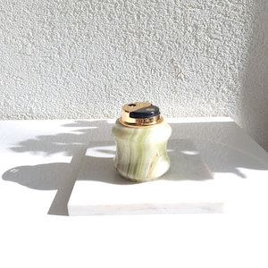 May include: A green and gold marble cigarette lighter with a black top. The lighter is sitting on a white marble surface.