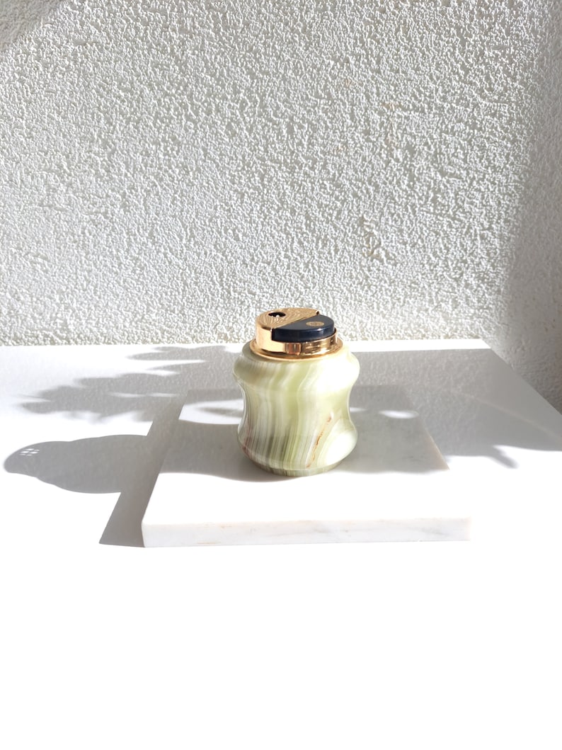 May include: A green and gold marble cigarette lighter with a black top. The lighter is sitting on a white marble surface.