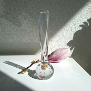 Op de afbeelding: Een heldere glazen vaas met een ronde, met bellen gevulde basis en een slanke hals. Een roze magnoliabloem en een kleine tak met knoppen liggen naast de vaas. De vaas heeft een klein label op de basis.