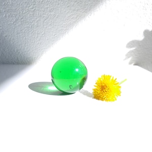 Op de afbeelding: Een groene glazen bol en een gele paardenbloem op een wit oppervlak.