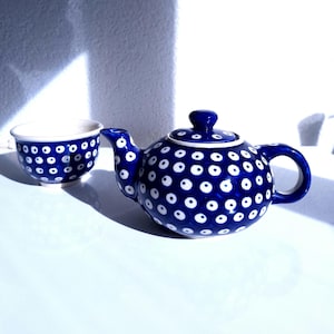 Op de afbeelding: Een keramische theepot en een bijpassend theekopje met een diepblauw glazuur, versierd met een herhalend patroon van witte cirkels met donkerblauwe centra. De theepot heeft een ronde vorm, een gebogen tuit, een handvat en een afneembaar deksel.