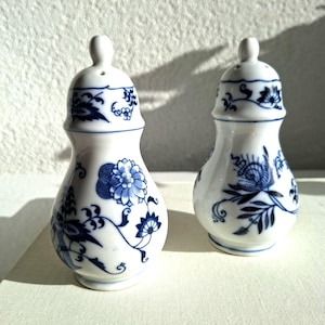 Op de afbeelding: Twee witte keramische zout- en peperstrooiers met blauwe bloemmotieven. Elke strooier heeft een afgeronde vorm, een kleine knop aan de bovenkant en kleine gaatjes. De blauwe patronen omvatten bloemen, bladeren en ranken.