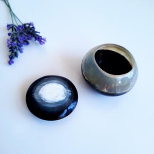 Op de afbeelding: Een kleine, ronde houder van gepolijst hoorn met een bijpassend deksel. De houder heeft een doorschijnend, rookgrijs bovendeel en een effen zwarte basis. Het deksel heeft een cirkelvormig, wit gemarmerd patroon in het midden, omringd door zwart.