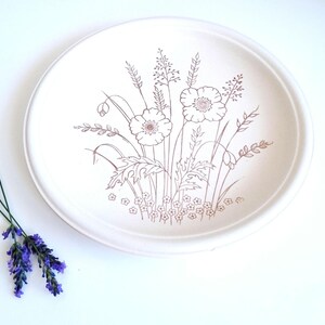 Op de afbeelding: Een rond wit keramieken bord met een bruine lijntekening van een boeket wilde bloemen met klaprozen en korenaren. Naast het bord liggen twee takjes paarse lavendel met groene stelen, wat een natuurlijk contrast vormt met het ontwerp.