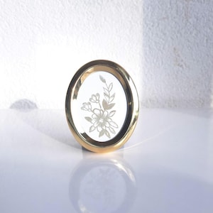 Può includere: Una cornice ovale dorata con un motivo floreale. La cornice ha un bordo dorato e uno sfondo bianco con un delicato motivo di fiori e foglie incise. La cornice è in piedi.