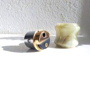 May include: A black and gold lighter with a round, textured body and a gold top. The lighter is sitting on a white surface next to a green and white marble container.