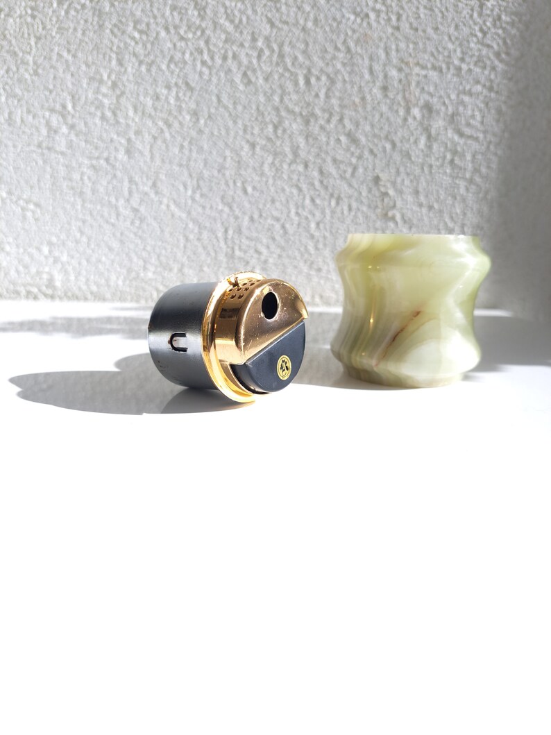 May include: A black and gold lighter with a round, textured body and a gold top. The lighter is sitting on a white surface next to a green and white marble container.