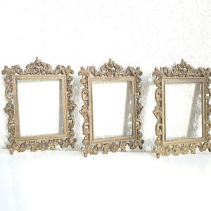 Puede incluir: Tres marcos de fotos ornamentados de estilo antiguo con acabado dorado. Cada marco tiene una abertura cuadrada y está decorado con intrincados adornos y motivos florales. Los marcos se muestran sobre una superficie blanca sobre un fondo blanco.