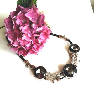 Op de afbeelding: Een ketting met een zwart koord, zilverkleurige metalen accenten en grote donkergrijze gefacetteerde kristallen. Het sieraad bevat kleine heldere strass-steentjes in de metalen schakels, gerangschikt in een boog naast een roze hortensiabloem.
