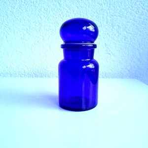 Op de afbeelding: Een kobaltblauwe glazen apothekerspot met een afgeronde stopper. De pot heeft een cilindrisch lichaam en een bijpassende stopper. De pot staat op een wit oppervlak tegen een lichtblauwe, getextureerde achtergrond.