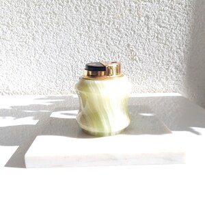 May include: A green and gold marble lighter with a black top. The lighter is sitting on a white marble surface.