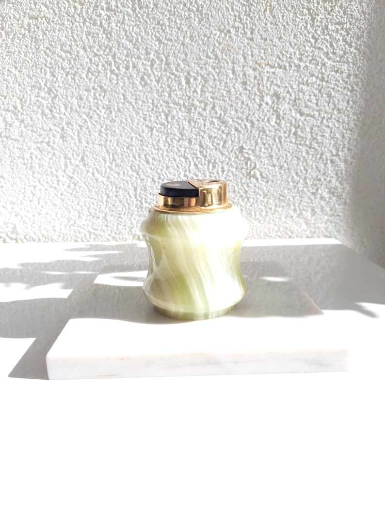 May include: A green and gold marble lighter with a black top. The lighter is sitting on a white marble surface.