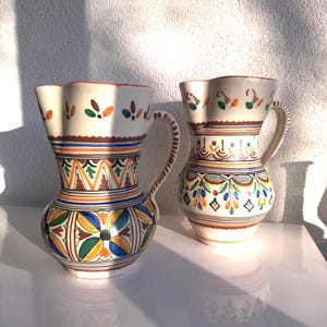 Op de afbeelding: Twee keramische kruiken met ingewikkelde, kleurrijke bloemen- en geometrische patronen. De kruiken hebben een crèmekleurige basis, met banden in oranje, blauw en groen. Elke kruik heeft een handvat en een geschulpte rand. De kruiken staan op een wit oppervlak.