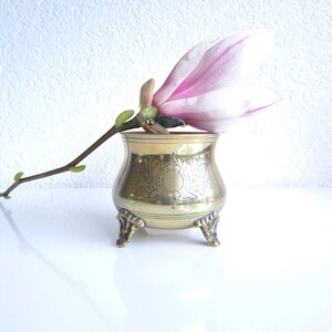 Peut inclure: Un petit vase à pied doré avec un motif orné. Une seule fleur de magnolia rose est placée dans le vase.