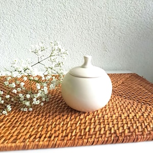 Op de afbeelding: Een witte keramische pot met een deksel, die op een geweven bruine placemat staat. Op de achtergrond is een takje met witte bloemen te zien.