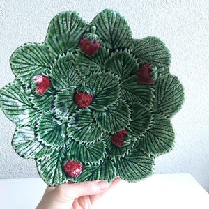 Peut inclure: Une assiette décorative en céramique avec un motif de fraises. L'assiette présente un bord festonné et est ornée de feuilles vertes et de fraises rouges. L'assiette est tenue par une main.