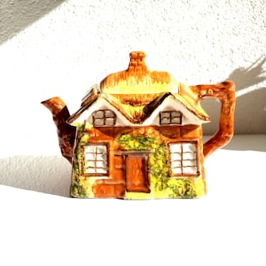 Può includere: Una teiera in ceramica marrone e gialla a forma di cottage con tetto di paglia, finestre e una porta. La teiera ha dei rampicanti di edera verdi dipinti sul lato.