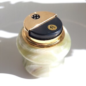 May include: A green marble cigarette lighter with a gold top and a black lid. The lid has a small gold emblem with a design.
