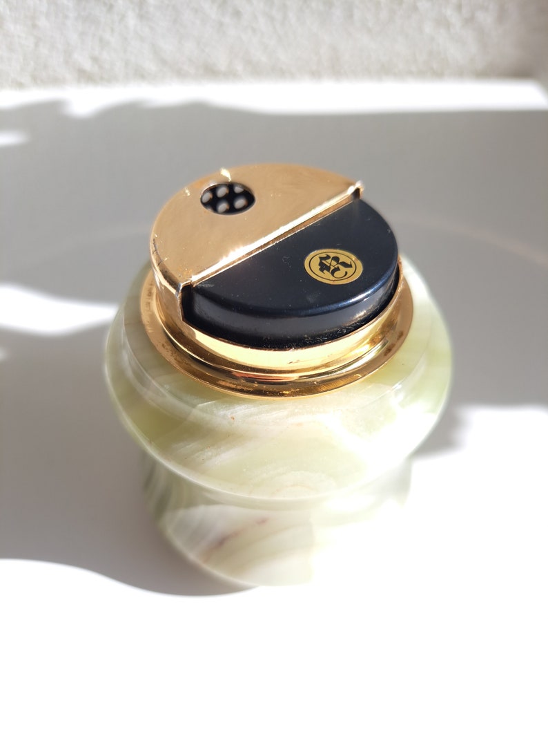 May include: A green marble cigarette lighter with a gold top and a black lid. The lid has a small gold emblem with a design.