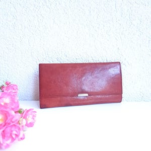 Puede incluir: Una cartera de cuero marrón rojizo con un cierre plateado. La cartera rectangular está cerrada. Hay rosas rosas en la esquina inferior izquierda y el fondo es una pared blanca texturizada.